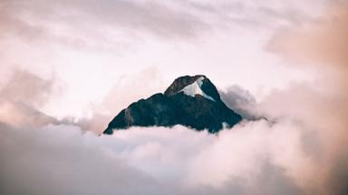 A dark mountain peak with a patch of snow rising above pink clouds. Photo: Cédric Servay via Wikimedia Commons, CC0 1.0 (cropped)