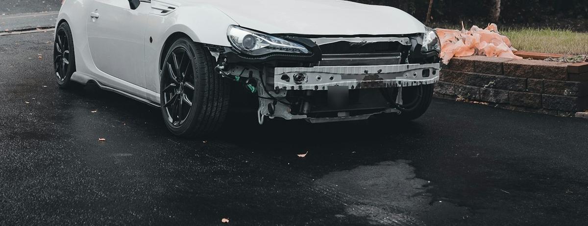 Front of a white car with a damaged bumper parked on a wet road. Photo: Erik Mclean on Unsplash (cropped)