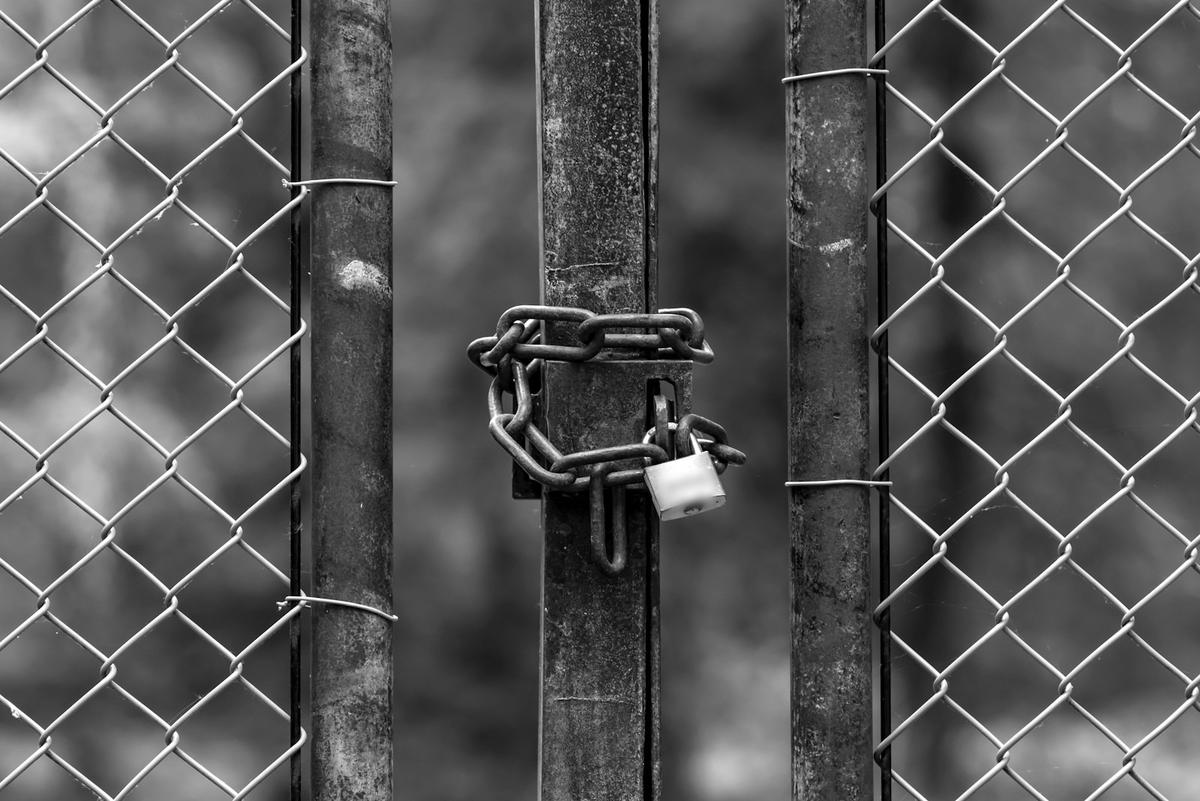 Chained and padlocked gate between two chain link fence panels, in black and white (edited). Photo: Lucas van Oort on Unsplash