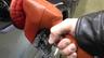 A bare hand in a leather jacket sleeve gripping an orange self-service fuel nozzle inserted in a silver car's fuel filler. Photo: Pumping gas by hand, MarkBuckawicki / Wikimedia Commons, CC0 (cropped and resized)