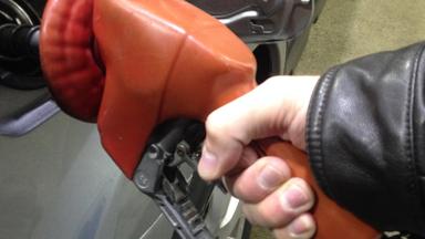 A bare hand in a leather jacket sleeve gripping an orange self-service fuel nozzle inserted in a silver car's fuel filler. Photo: Pumping gas by hand, MarkBuckawicki / Wikimedia Commons, CC0 (cropped and resized)