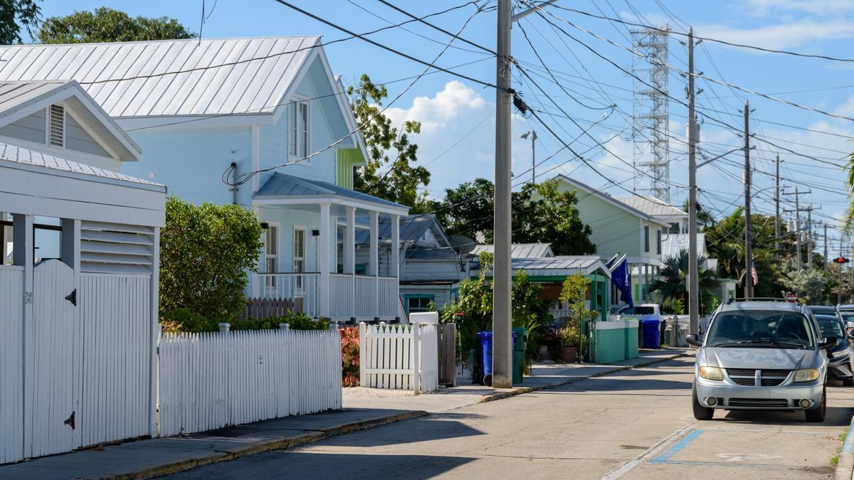 A quiet residential street in Key West, Florida, lined with pastel wooden houses, white picket fences and palm trees under a blue sky. Photo: Residential Street in Key West, Florida, Julian Lupyan / Wikimedia Commons, CC0 (cropped and resized)
