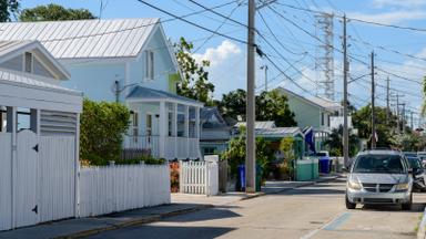 A quiet residential street in Key West, Florida, lined with pastel wooden houses, white picket fences and palm trees under a blue sky. Photo: Residential Street in Key West, Florida, Julian Lupyan / Wikimedia Commons, CC0 (cropped and resized)