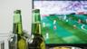 A flat-screen TV showing a blurred football match behind two unlabeled green glass bottles and a drinking glass on a table. Photo: jeshoots.com / Wikimedia Commons, CC0 (cropped and resized)