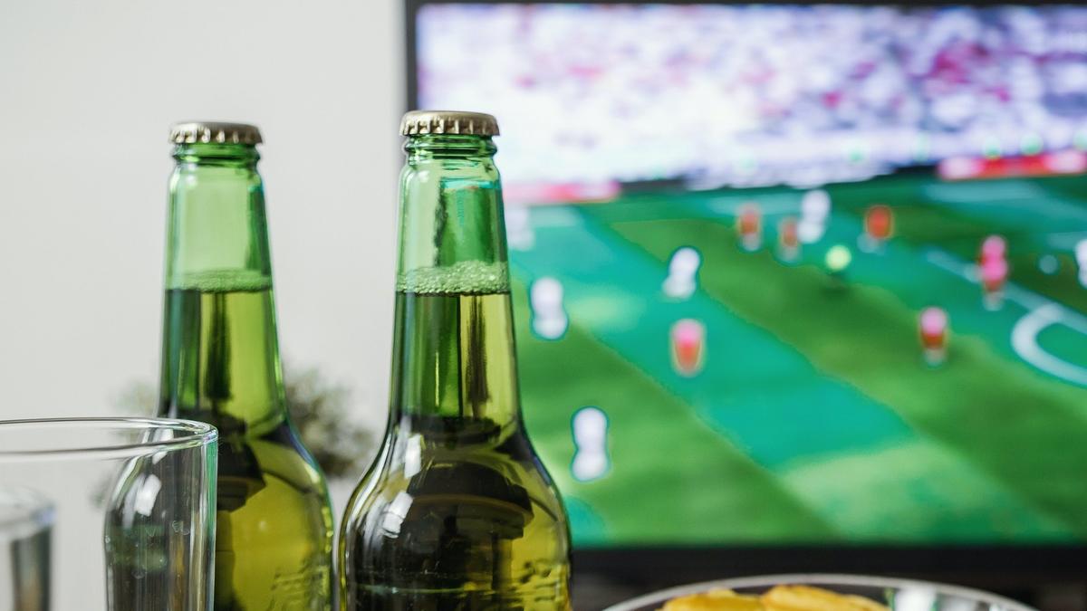 A flat-screen TV showing a blurred football match behind two unlabeled green glass bottles and a drinking glass on a table. Photo: jeshoots.com / Wikimedia Commons, CC0 (cropped and resized)