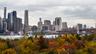 Lower Manhattan skyline and the Brooklyn Bridge tower seen across the East River past autumn trees under an overcast sky. Photo: Financial District East Skyline, 2025, Julian Lupyan / Wikimedia Commons, CC0 (cropped and resized)
