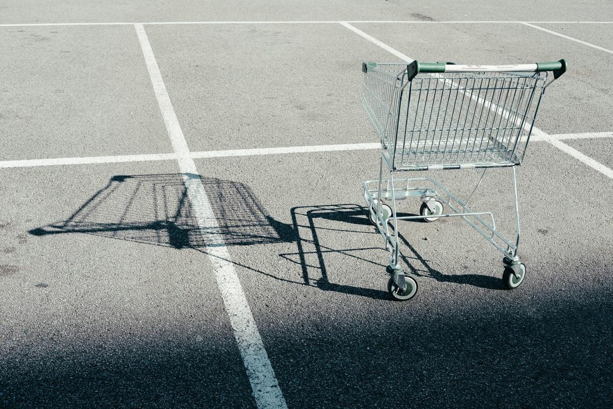 A gray metal shopping cart and its shadow on an empty parking lot with white painted lines. Photo: David Clarke on Unsplash.