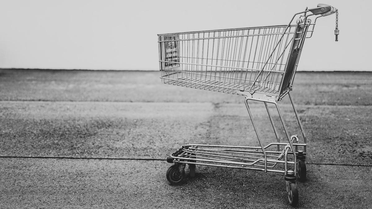 An empty metal shopping cart standing on an open stretch of pavement in black and white. Photo: Bruno Kelzer via Unsplash
