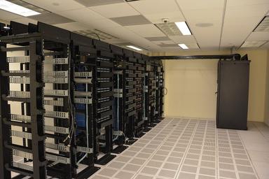 Rows of open black server racks with network cabling on a raised data center floor. Photo: Carl Lender / Wikimedia Commons, CC BY 2.0